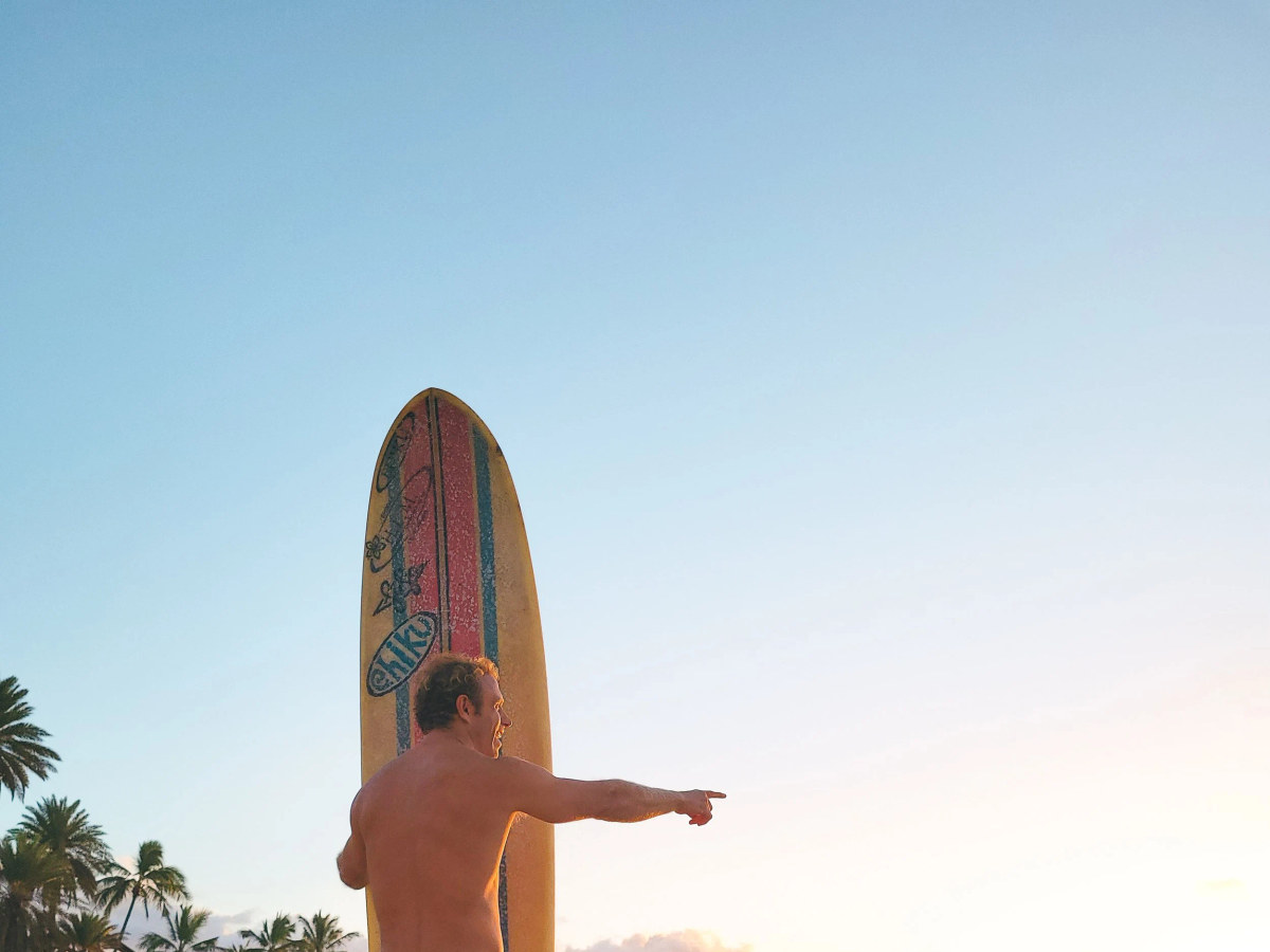 An SFC surfer holding a longboard on the beach at sunset in Hawaiʻi